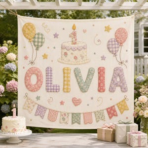 May include: A cream-colored fabric wall hanging featuring a quilted birthday cake with a single candle, balloons, and a bunting banner. The name OLIVIA is appliqued in large, floral and gingham patterned letters across the center of the textile.
