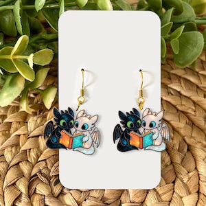 May include: A pair of dangle earrings featuring a black and white dragon couple reading a book. The earrings have gold-colored hooks and are set against a white background. The dragons are holding a book with orange and blue pages.
