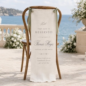 May include: A wooden chair draped with a cream-colored fabric sign featuring sage green ribbons. The text reads: This seat is reserved in loving memory of Thomas Hayes, father of the bride. Forever loved, forever remembered, forever part of our day.