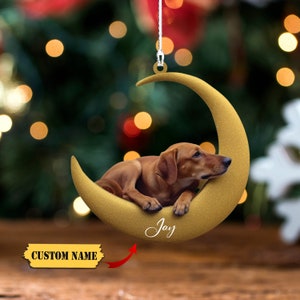 Puede incluir: Adorno navideño con un perro salchicha marrón descansando en una luna creciente dorada. El adorno tiene el nombre "Jay" escrito. Las palabras "CUSTOM NAME" están en una pancarta amarilla.