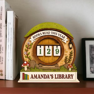 May include: Wooden book counter with a hobbit-hole design. The counter displays "129" and the text "BOOKS READ THIS YEAR". The base reads "AMANDA'S LIBRARY". The design includes a green roof, mushrooms, and floral accents.