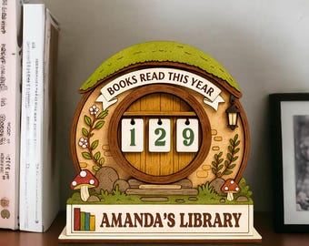 May include: Wooden book counter with a hobbit-hole design. The counter displays "129" and the text "BOOKS READ THIS YEAR". The base reads "AMANDA'S LIBRARY". The design includes a green roof, mushrooms, and floral accents.