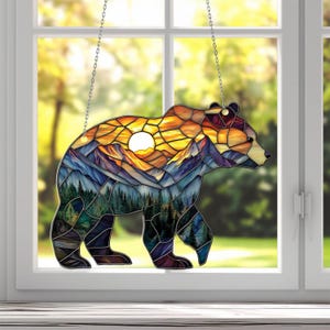 Puede incluir: Una ventana de vidrio coloreado que cuelga con una silueta de oso. El cuerpo del oso está lleno de una escena de montaña colorida con una puesta de sol y un sol amarillo brillante.