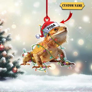 May include: A Christmas ornament featuring a bearded dragon wearing a Santa hat with the name "YODA" and wrapped in colorful Christmas lights. The ornament hangs from a red ribbon, with a snowy background and a Christmas tree.