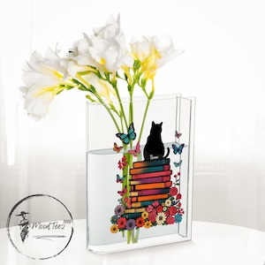 May include: A clear glass vase with a black cat silhouette sitting on a stack of colorful books. The vase is filled with water and white flowers. The design includes butterflies and flowers.
