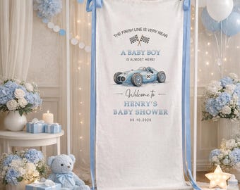 May include: A baby shower backdrop with a light blue race car illustration and the text "A Baby Boy is Almost Here!" The backdrop is decorated with blue ribbons and balloons. The text "Welcome to Henry's Baby Shower 05.10.2026" is also visible.