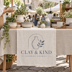 May include: A beige linen table runner featuring a dark blue logo with an arched outline, a vase, and a leafy branch, with the text CLAY & KIND HANDMADE CERAMICS printed below. The runner is displayed on a table filled with various ceramic bowls and vases.