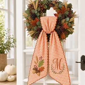Puede incluir: Una corona decorativa de oto&ntilde;o con hojas de eucalipto verdes, pi&ntilde;as marrones, tallos de trigo y hojas de arce naranjas. Una cinta de tela de cuadros vichy naranja y blanca cuelga del centro, bordada con dos bellotas y la letra M en cursiva.