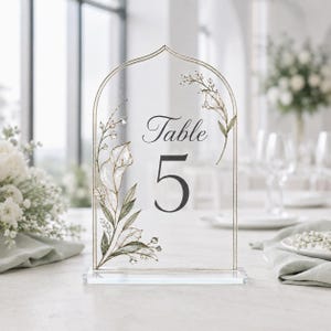 May include: An arched clear acrylic table number sign featuring the text Table 5 in a dark gray serif font. The sign is decorated with delicate botanical illustrations of white calla lilies and green leaves with gold outlines on the sides.