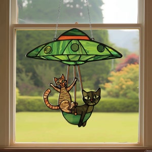 May include: A stained glass window hanging features two cats riding in a green flying saucer. The cats are brown and gray with black eyes. The saucer has a red stripe around the top.