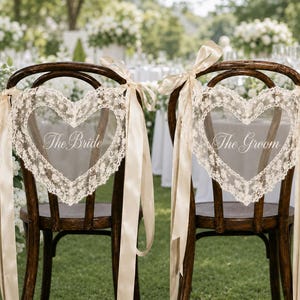 May include: Two dark wood chairs featuring heart-shaped lace back covers with long champagne-colored satin ribbons tied at the sides. The left chair has the text The Bride embroidered in white, and the right chair has the text The Groom embroidered in white.