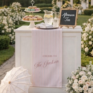 May include: An outdoor afternoon tea setup with a white table, a tiered dessert stand with macarons, and a sign that says "Afternoon Tea." A pink and white striped runner reads "Cheers to The Jackson."
