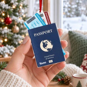 May include: A flat, rectangular ornament shaped like a blue United States passport with a globe icon, the text PASSPORT, United States of America, and Name. A light blue boarding pass with the text BOARDING PASS, Name, From, and Destination is tucked inside.