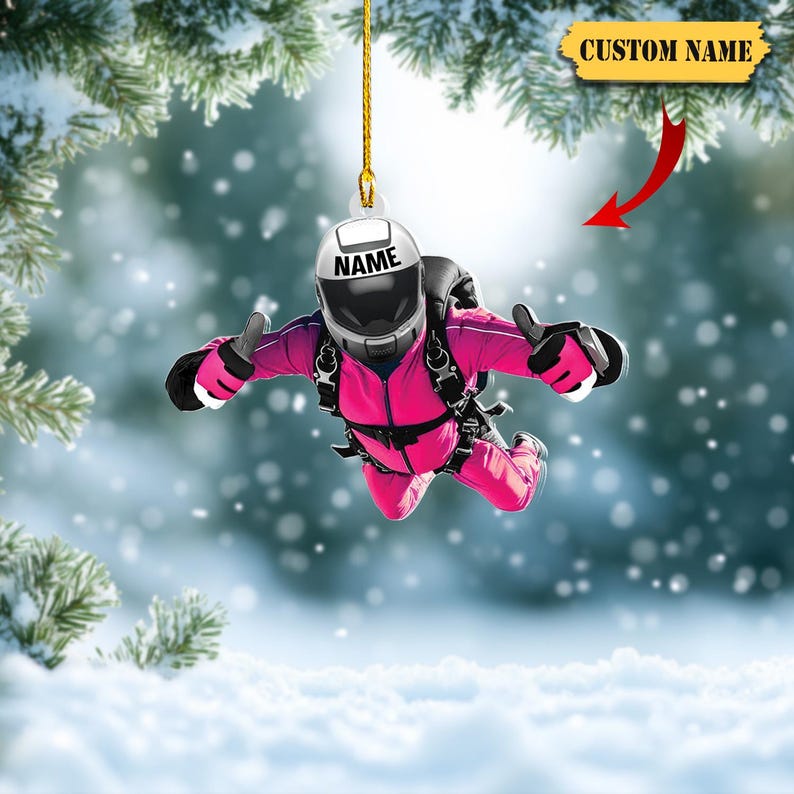 Pu&ograve; includere: Un ornamento rosa per paracadutismo con casco e attrezzatura neri. Il casco ha la scritta "NAME". L'ornamento &egrave; appeso a un filo d'oro. Un banner in alto dice "CUSTOM NAME".