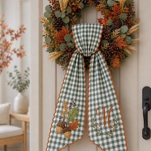 Puede incluir: Una corona de oto&ntilde;o con pi&ntilde;as, trigo y hojas naranjas, adornada con un colgante de tela de cuadros verdes y blancos. La tela presenta bellotas y trigo bordados en un lado y una letra M monogramada en el otro lado.