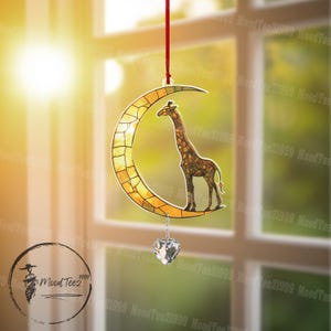 Puede incluir: Un atrapasol de vidrio tintado con una luna creciente con una jirafa de pie sobre ella. El atrapasol está colgado de una cuerda roja y tiene un corazón de cristal colgando de la parte inferior.