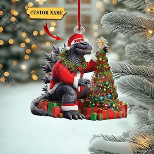 May include: A Christmas ornament featuring Godzilla in a Santa Claus outfit, decorating a miniature Christmas tree. The ornament is black, red, and green, with a custom name tag. Presents are at the base of the tree.