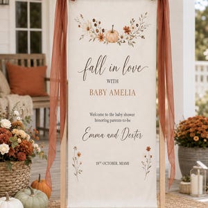 May include: A vertical fabric banner on a wooden stand with rust-colored chiffon bows. The banner features a floral pumpkin illustration and text reading: fall in love WITH BABY AMELIA Welcome to the baby shower honoring parents-to-be Emma and Dexter 19TH OCTOBER, MIAMI.