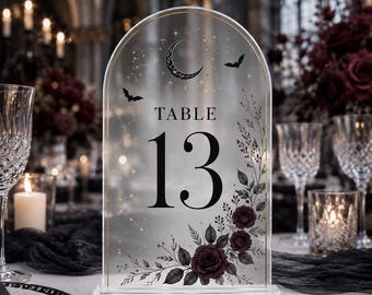 Könnte beinhalten: Ein bogenförmiges Tischaufstellerschild aus klarem Acryl mit der schwarzen Aufschrift TABLE 13. Das Design zeigt einen Halbmond, Sterne, Sternbilder, zwei fliegende Fledermäuse und ein Arrangement aus dunkelroten Rosen mit schwarzen Blättern.