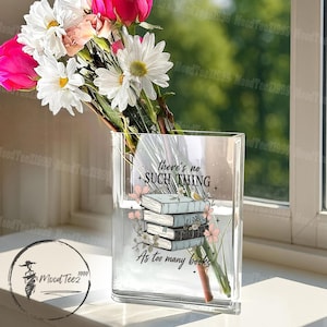 May include: A clear glass vase with a floral print design featuring a stack of books and the text "there's no such thing as too many books". The vase is filled with white and pink flowers.