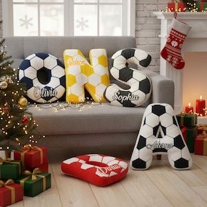 May include: Decorative soccer ball-themed pillows shaped as the letters O, N, S, and A, with names embroidered on them. The letter O is navy blue, N is yellow, S is black and white, and A is black and white. A red and white pillow is also present.