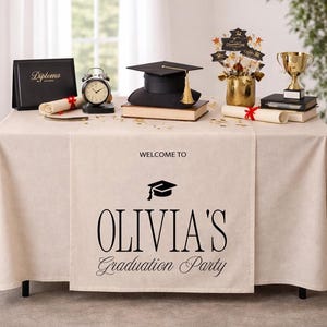 May include: A graduation party table setting with a black mortarboard, diploma, trophy, and clock. The table runner reads "Welcome to Olivia's Graduation Party". Gold and black decorations are scattered around the table, creating a celebratory atmosphere.