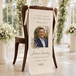 May include: A cream-colored fabric memorial sign draped over a dark wooden chair, featuring the text A SEAT SAVED IN YOUR HONOR, Always with us, James Sanchez, FATHER OF THE BRIDE, October 13, 2026, and a portrait of a man in a blue suit.