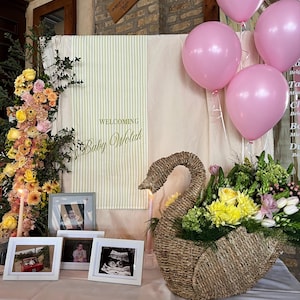 May include: A baby shower display featuring a woven swan basket filled with yellow and green flowers. Pink balloons float above a table with framed photos and a sign that reads "Welcoming Baby Wolah."