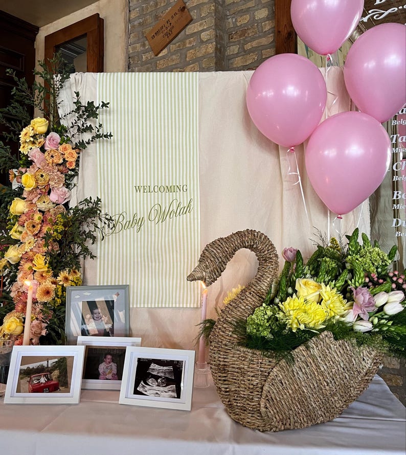 May include: A baby shower display featuring a woven swan basket filled with yellow and green flowers. Pink balloons float above a table with framed photos and a sign that reads "Welcoming Baby Wolah."
