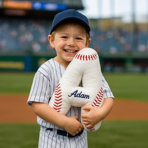 May include: A child in a baseball uniform and navy cap smiles, holding a large, white baseball-style pillow shaped like the letter "A". The pillow features red stitching and the name "Adam" in blue, a fun, personalised gift.