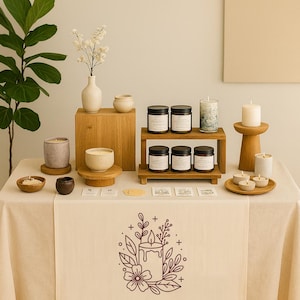 May include: A display of candles and home decor items on a table covered with a cream-colored tablecloth. The table features various candles, wooden stands, and small decorative bowls. The tablecloth has a floral candle logo.