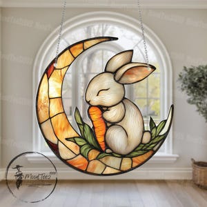 以下が含まれることがあります： 吊り下げられたステンドグラスの窓には、三日月とそこに座っている白いウサギが描かれています。ウサギは足にニンジンを持っています。月は黄色、オレンジ、茶色のステンドグラスで飾られています。