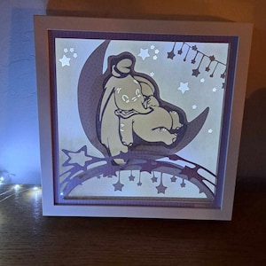 Peut inclure: Une boîte à lumière encadrée de blanc avec une lumière à l'intérieur. La boîte à lumière présente un motif découpé au papier de Dumbo l'éléphant assis sur un croissant de lune avec des étoiles en arrière-plan.