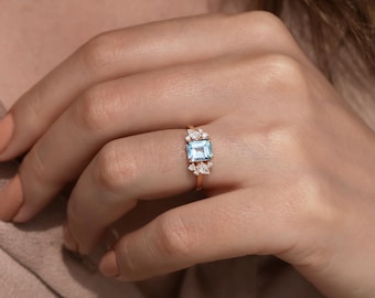 May include: A rose gold ring with a light blue square-shaped gemstone surrounded by smaller clear gemstones.
