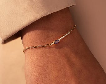 Könnte beinhalten: Ein goldenes Kettenarmband mit einem blauen Edelstein und kleinen klaren Steinen.