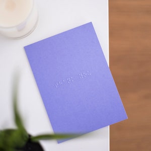 A vibrant purple card lay flat on a table with thank you written in lower case grade one braille. There is a candle in the top left and a blurry plant in the bottom left.