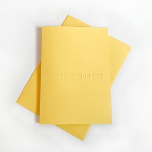 Image shows a blue card with the words happy birthday written in grade 1 UEB braille. A matching yellow envelope is beneath the card.