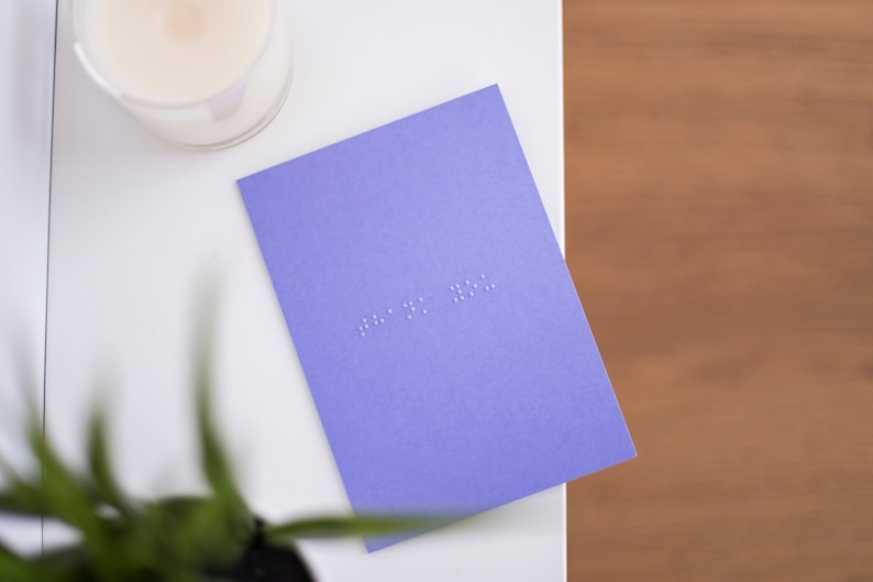 A vibrant purple card lay flat on a table with thank you written in lower case grade one braille. There is a candle in the top left and a blurry plant in the bottom left.