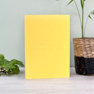 A vibrant yellow card with happy father's day written in lower case braille. There is foliage to the left of the card and a plant on the right.