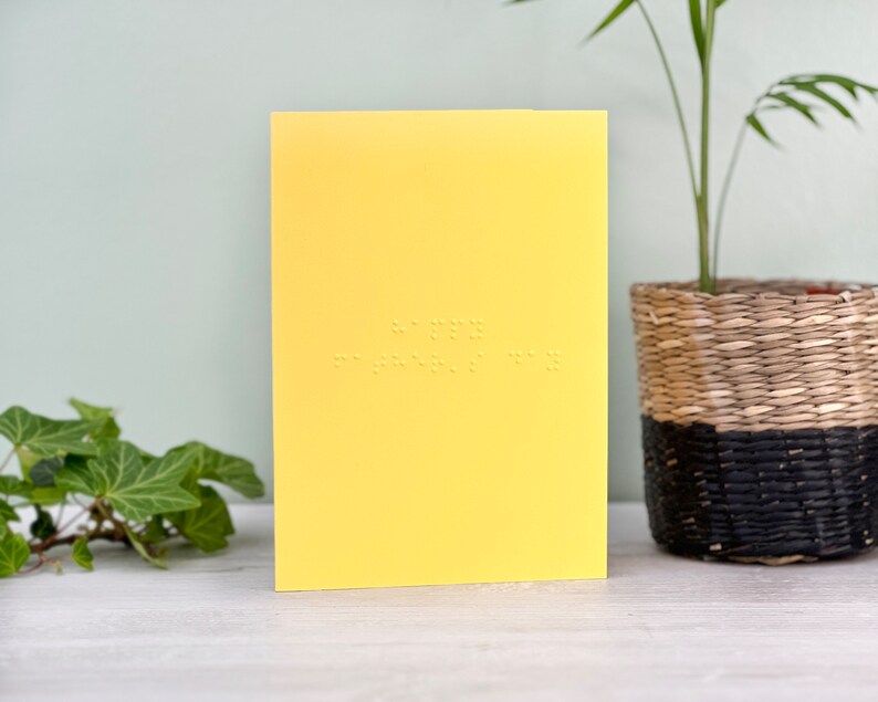 A vibrant yellow card with happy father's day written in lower case braille. There is foliage to the left of the card and a plant on the right.