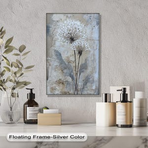 a floating frame-silver color artwork hanging on a wall, with a white vase of flowers and various toiletries, including bottles and a pump, arranged on a marble countertop below the frame.