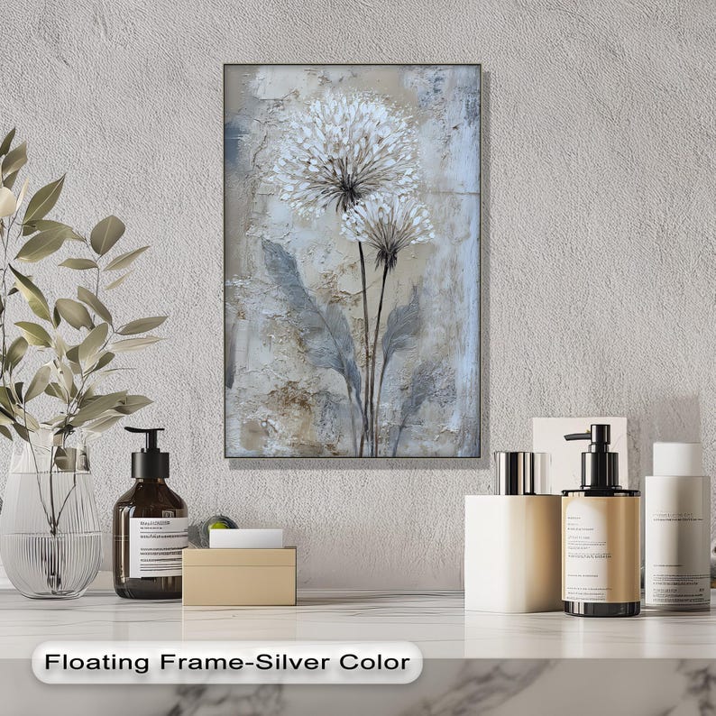 a floating frame-silver color artwork hanging on a wall, with a white vase of flowers and various toiletries, including bottles and a pump, arranged on a marble countertop below the frame.