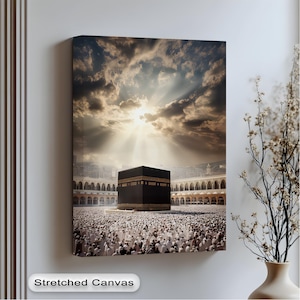 Könnte beinhalten: Aufgespanntes Leinwandbild, das die Kaaba in Mekka, Saudi-Arabien, unter einem dramatischen Himmel mit Sonnenstrahlen zeigt. Die schwarze, würfelförmige Struktur ist von einer großen Menschenmenge in weißen Gewändern umgeben. Die Leinwand ist auf einen Holzrahmen gespannt.