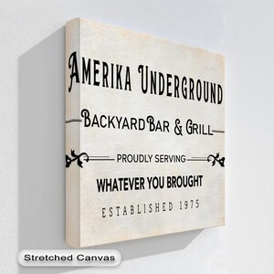 May include: A stretched canvas wall art piece with the text "AMERIKA UNDERGROUND BACKYARD BAR & GRILL" in black. The text "PROUDLY SERVING WHATEVER YOU BROUGHT ESTABLISHED 1975" is also displayed. The canvas has a vintage, off-white background.