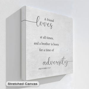 Op de afbeelding: Witte canvas wall art met de tekst "A friend loves at all times, and a brother is born for a time of adversity" en "Proverbs 17:17". De woorden zijn in een zwart script.