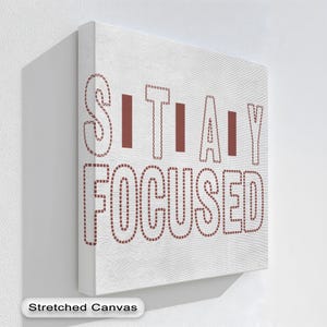 以下が含まれることがあります： 「STAY FOCUSED」の文字がステッチ風のフォントで描かれた、白いストレッチキャンバスのウォールアート。文字は赤茶色の点線模様で縁取られ、背景は白です。