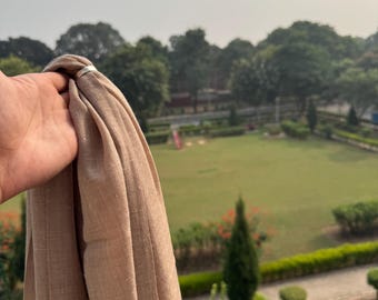 May include: A light brown scarf made of a soft, woven fabric. The scarf is held in a hand and is draped over a railing.