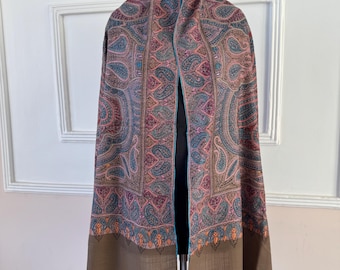 May include: A patterned shawl in pink, blue, and brown hues, featuring a detailed paisley design. The shawl is displayed on a form, highlighting its intricate pattern and fringed edges. The bottom border is a solid brown.