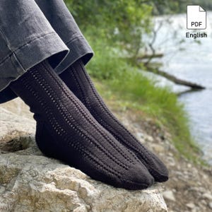 Könnte beinhalten: Nahaufnahme von zwei schwarzen Stricksocken mit einem strukturierten Lochmuster. Die Socken werden mit grauen Hosen getragen und liegen auf einem hellen Felsen. Der Hintergrund zeigt Wasser und grüne Vegetation.