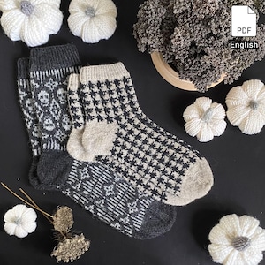 May include: Two pairs of knitted socks with patterned designs. One pair features a skull and floral pattern, the other a houndstooth pattern. The socks are in shades of grey, black, and beige. The image also includes decorative white knitted pumpkins and a pot of dried lavender.